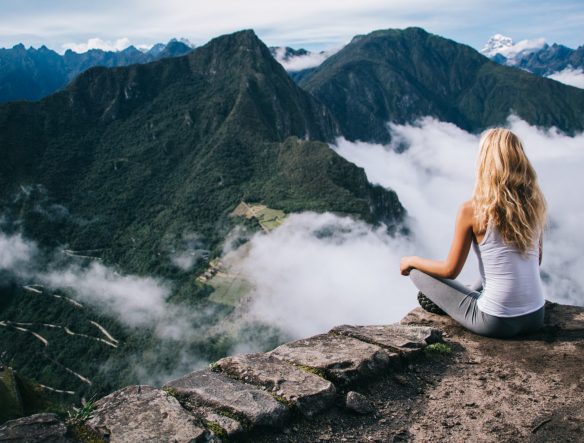 Back view of woman tourist sitting in lotus pose relaxing and admiring breathtaking view during trek tour, female traveler enjoying beauty of Machu Picchu landscape and high mountains covered clouds